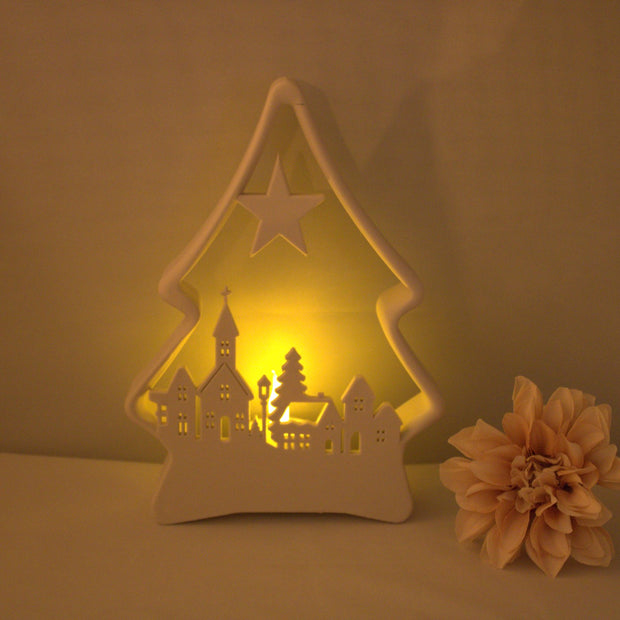 Witte kunststof kerstboom verlicht met warm geel LED waxinelichtje en winterdorp silhouet – sfeervolle kerstdecoratie

